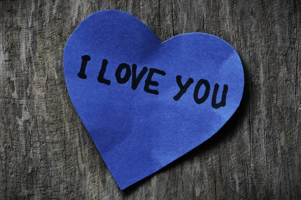 Seni mavi kalp şeklindeki kağıt kartta seviyorum. Tahta ahşap kaplamalı arkaplanda sevgililer günü için. Sevgililer günü kartında seni seviyorum yazıyor. Minimalist Sevgililer Günü.