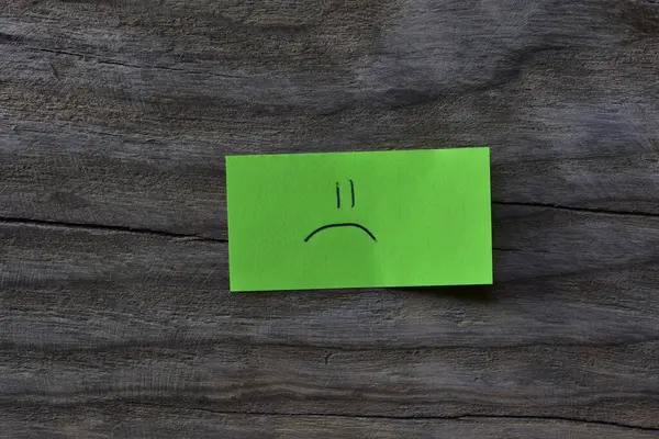 Ahşap kaplamalı arka planda yeşil yapışkan nottaki üzgün surat. Not kağıdına üzgün bir surat çiz..