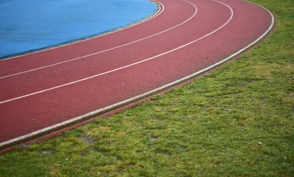 Sol tarafta mavi spor sahası olan kırmızı koşu pisti ve sağ tarafta yeşil çimenler, güneşli bir gün.