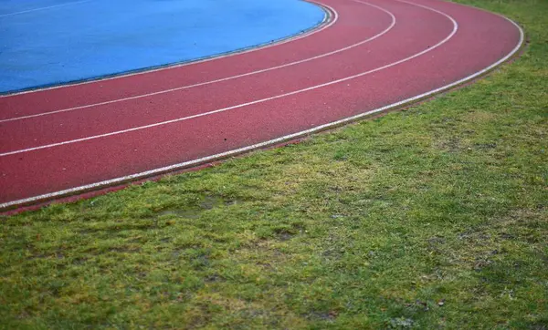 Sol tarafta mavi spor sahası olan kırmızı koşu pisti ve sağ tarafta yeşil çimenler, güneşli bir gün.