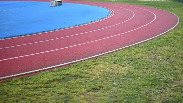 Sol tarafta mavi spor sahası olan kırmızı koşu pisti ve sağ tarafta yeşil çimenler, güneşli bir gün.