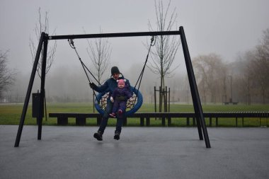 Baba, kızıyla birlikte sonbaharda sisli bir günün dışında çocuk parkında sallanıyor. Baba ve kızı birlikte vakit geçiriyorlar..