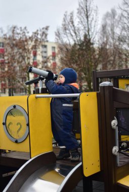 Küçük çocuk soğuk kış zamanı halka açık parkta oyuncak teleskopla oynuyor. Çocuk oyun alanında teleskop kullanıyor.