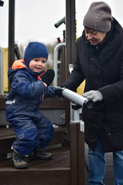 Küçük çocuk soğuk kış zamanında halka açık parkta oyuncak teleskopla oynuyor. Büyükannem torununa teleskobu nasıl kullanacağını öğretiyor. İki nesil dışarıda zaman geçiriyor.