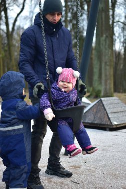 Babam, kış günü parkta salıncakta sallanan bir kızı sallıyor. Küçük kardeş onların yanında duruyor. Dışarıda eğlenceli vakit geçiriyoruz.