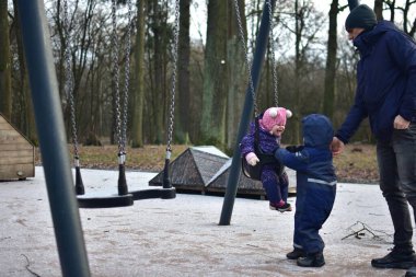 Babam, kış günü parkta salıncakta sallanan bir kızı sallıyor. Küçük kardeş onların yanında duruyor. Dışarıda eğlenceli vakit geçiriyoruz.