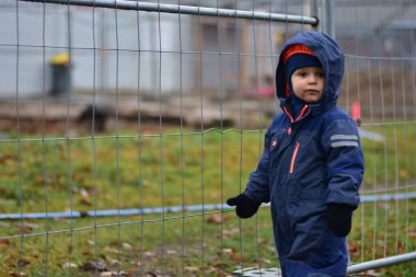 Sonbaharda şantiyeyi gözetleyen kapüşonlu meraklı bir çocuk. Yeni yürümeye başlayan çocuk çitin yanında duruyor. Çocuk inşaatın nasıl işlediğini izliyor.