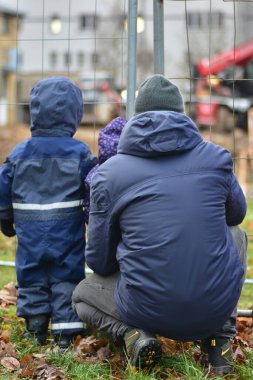 Küçük çocuk, kız ve babaları çitten şantiyeye bakıyor. Meraklı çocuklar ve babalar inşaatın nasıl işlediğini izliyor.