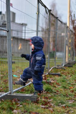 Sonbaharda şantiyeyi gözetleyen kapüşonlu meraklı bir çocuk. Yeni yürümeye başlayan çocuk çitin yanında duruyor. Çocuk inşaatın nasıl işlediğini izliyor.