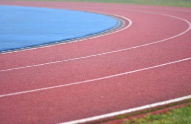 Beyaz çizgileri olan kırmızı koşu yolu eğrisi, sol tarafta mavi spor sahası ve sağ tarafta yeşil çimen
