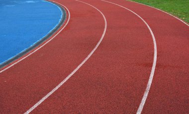Beyaz çizgileri olan kırmızı koşu yolu eğrisi, sol tarafta mavi spor sahası ve sağ tarafta yeşil çimen