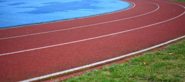 Beyaz çizgileri olan kırmızı koşu yolu eğrisi, sol tarafta mavi spor sahası ve sağ tarafta yeşil çimen