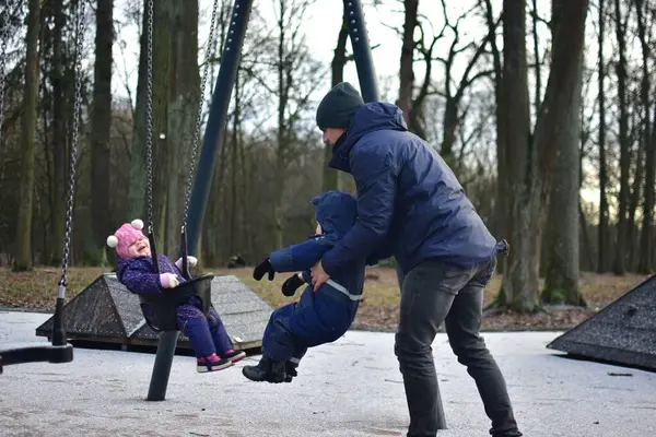 Babam, kış günü parkta salıncakta sallanan bir kızı sallıyor. Küçük kardeş onların yanında duruyor. Dışarıda eğlenceli vakit geçiriyoruz.