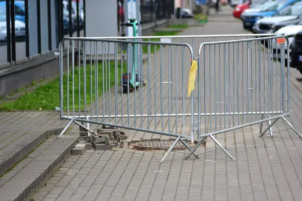 Güvenlik bariyeri hasarlı bir kaldırımı, gevşek tuğlaları ve şehir ortamında açık bir rögar kapağı ile çevreler. Yaya kazalarını önlemek için inşaat veya onarım alanı işaretlendi.