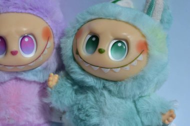 Kabarık mavi ve pembe canavar kostümlü iki oyuncu Labubu oyuncak figürü ve doğrudan güneş ışığı altında beyaz bir arkaplan karşısında muzip bir gülümseme..