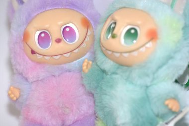 Kabarık yeşil ve pembe canavar kostümlü iki oyuncu Labubu oyuncak figürü ve beyaz bir arka plana karşı muzip bir gülümseme..