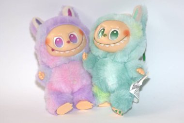 Kabarık yeşil ve pembe canavar kostümlü iki oyuncu Labubu oyuncak figürü ve beyaz bir arka plana karşı muzip bir gülümseme..