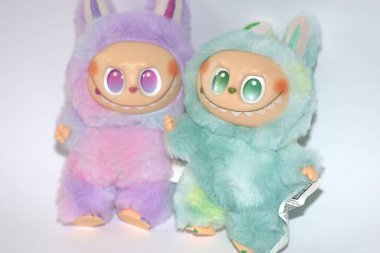 Kabarık yeşil ve pembe canavar kostümlü iki oyuncu Labubu oyuncak figürü ve beyaz bir arka plana karşı muzip bir gülümseme..