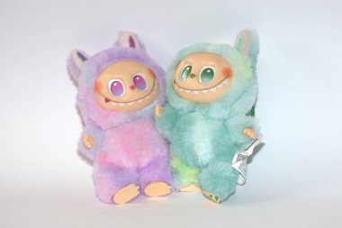 Kabarık yeşil ve pembe canavar kostümlü iki oyuncu Labubu oyuncak figürü ve beyaz bir arka plana karşı muzip bir gülümseme..