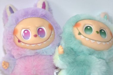 Kabarık yeşil ve pembe canavar kostümlü iki oyuncu Labubu oyuncak figürü ve beyaz bir arka plana karşı muzip bir gülümseme..
