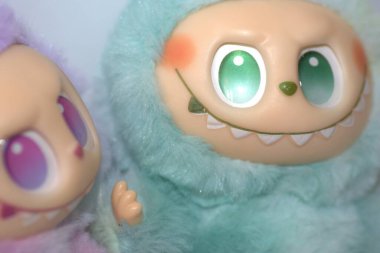 Kabarık yeşil ve pembe canavar kostümlü iki oyuncu Labubu oyuncak figürü ve beyaz bir arka plana karşı muzip bir gülümseme..