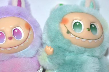 Kabarık yeşil ve pembe canavar kostümlü iki oyuncu Labubu oyuncak figürü ve beyaz bir arka plana karşı muzip bir gülümseme..