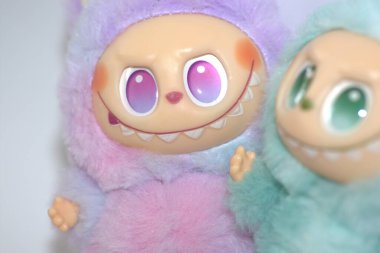 Kabarık yeşil ve pembe canavar kostümlü iki oyuncu Labubu oyuncak figürü ve beyaz bir arka plana karşı muzip bir gülümseme..