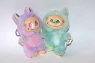 Kabarık yeşil ve pembe canavar kostümlü iki oyuncu Labubu oyuncak figürü ve beyaz bir arka plana karşı muzip bir gülümseme..
