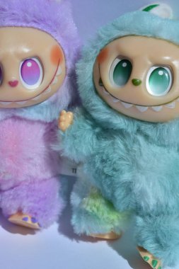 Kabarık mavi ve pembe canavar kostümlü iki oyuncu Labubu oyuncak figürü ve doğrudan güneş ışığı altında beyaz bir arkaplan karşısında muzip bir gülümseme..