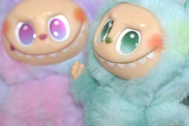 Kabarık yeşil ve pembe canavar kostümlü iki oyuncu Labubu oyuncak figürü ve beyaz bir arka plana karşı muzip bir gülümseme..