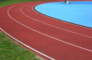 Kırmızı koşu parkuru sağ tarafta mavi spor sahası ve sol tarafta yeşil çimenler, güneşli bir gün.