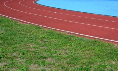 Kırmızı koşu parkuru sağ tarafta mavi spor sahası ve sol tarafta yeşil çimenler, güneşli bir gün.