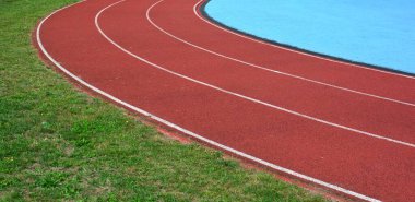 Kırmızı koşu parkuru sağ tarafta mavi spor sahası ve sol tarafta yeşil çimenler, güneşli bir gün.