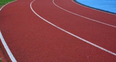 Kırmızı koşu parkuru sağ tarafta mavi spor sahası ve sol tarafta yeşil çimenler, güneşli bir gün.