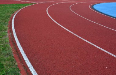 Kırmızı koşu parkuru sağ tarafta mavi spor sahası ve sol tarafta yeşil çimenler, güneşli bir gün.