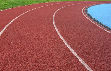 Kırmızı koşu parkuru sağ tarafta mavi spor sahası ve sol tarafta yeşil çimenler, güneşli bir gün.