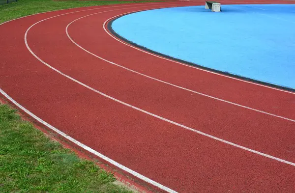 Kırmızı koşu parkuru sağ tarafta mavi spor sahası ve sol tarafta yeşil çimenler, güneşli bir gün.