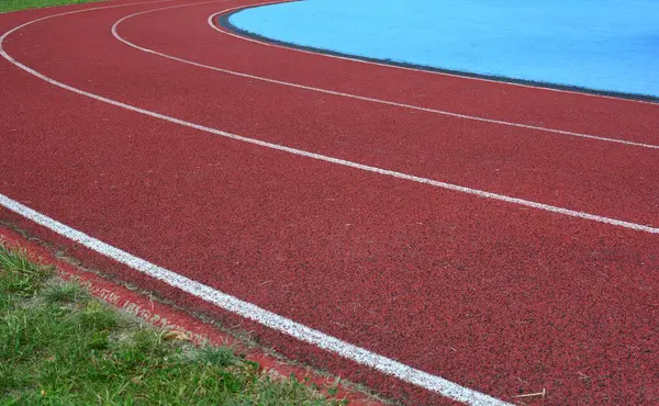 Kırmızı koşu parkuru sağ tarafta mavi spor sahası ve sol tarafta yeşil çimenler, güneşli bir gün.