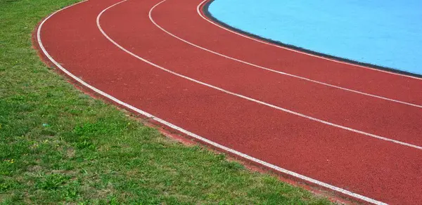Kırmızı koşu parkuru sağ tarafta mavi spor sahası ve sol tarafta yeşil çimenler, güneşli bir gün.