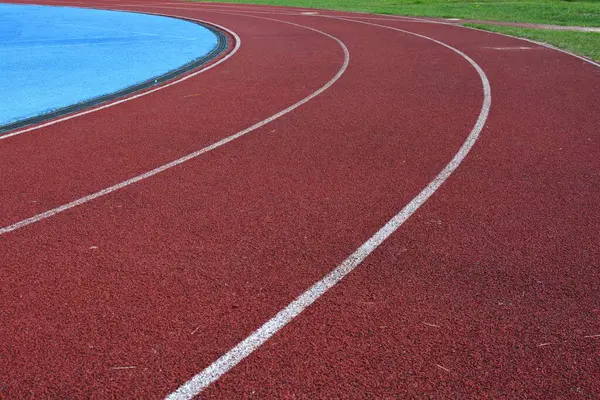 Beyaz çizgileri olan kırmızı koşu yolu eğrisi, sol tarafta mavi spor sahası ve sağ tarafta yeşil çimen