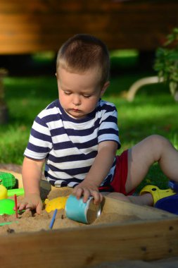Soyunmuş tişörtlü çocuk arka bahçede kum havuzunda oynuyor. Çocuk yaz mevsiminde dışarıda yoğunlaşır..