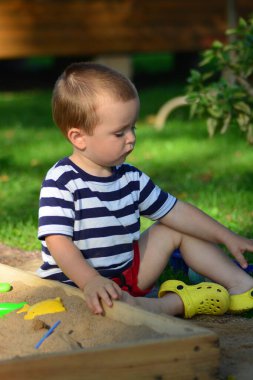 Sarı sandaletli çocuk arka bahçedeki kum havuzunda oynuyor. Çocuk, yaz mevsiminde dışarıya bakmaya odaklanmış..