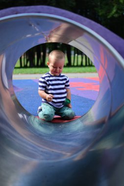 Küçük çocuk oyun parkında sürünme tünelinin diğer tarafında oturuyor. Çocuk oyun parkında sürünme tüneline giriyor. Yuvarlak çerçeveli çocuk portresi..
