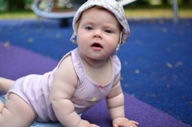 Küçük kız sıcak yaz günlerinde çocuk parkındaki kameraya bakıp sürünüyor. Bebek, çocuk parkında vakit geçiriyor..