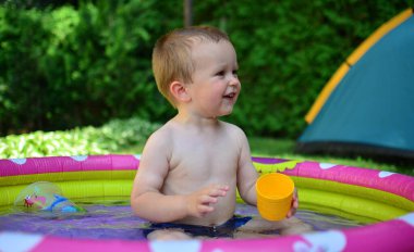 Küçük çocuk sıcak yaz gününde küçük bir su havuzunda suyla oynuyor. Çocuk dışarıdaki havuzda eğleniyor. Çocuk şişme havuzda oturuyor ve gülümsüyor..