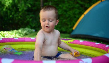 Küçük çocuk sıcak yaz gününde küçük bir su havuzunda suyla oynuyor. Çocuk dışarıdaki havuzda eğleniyor. Çocuk şişme havuzda oturuyor ve su sıçrıyor..