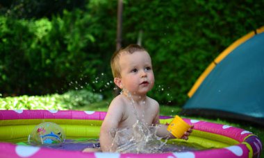 Küçük çocuk sıcak yaz gününde küçük bir su havuzunda suyla oynuyor. Çocuk dışarıdaki havuzda eğleniyor. Çocuk şişme havuzda oturuyor ve su sıçrıyor..