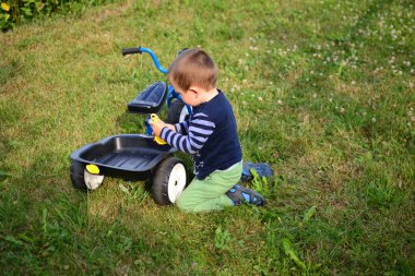 Küçük çocuk arka bahçede mavi üç tekerlekli bisikletin yanında oturuyor ve kum oyuncağını üç tekerlekli bisikletin bagajına koyuyor. Küçük çocuk yazın dışarıda üç tekerlekli bisikletle oynuyor..