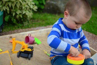 Küçük çocuk arka bahçedeki kum havuzunda oynuyor. Çocuk yazın dışarıda oyuncak tavayla kum yemeği yapıyor..