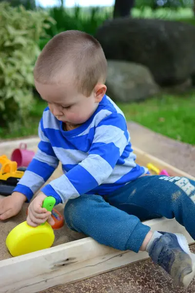 Küçük çocuk arka bahçedeki kum havuzunda oynuyor. Çocuk yazın dışarıda oyuncak tavayla kum yemeği yapıyor..